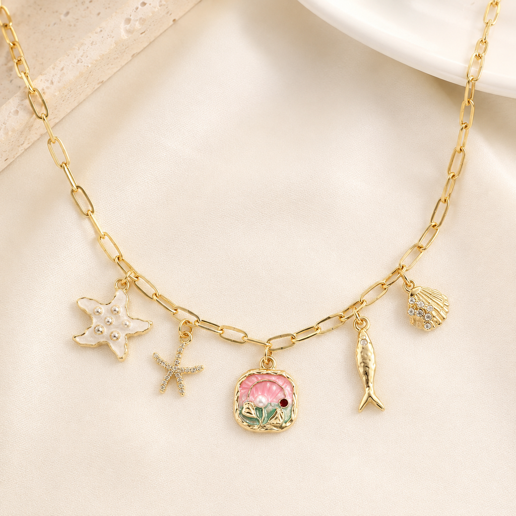 Gold chain necklace with multiple charms including a pink seashell, starfish, fish, and shell, displayed on a soft cream fabric background.