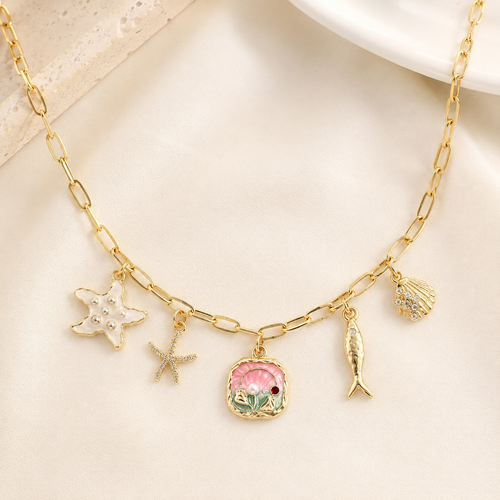 Gold chain necklace with multiple charms including a pink seashell, starfish, fish, and shell, displayed on a soft cream fabric background.