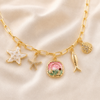 Gold paperclip chain necklace with sea-themed charms including a white pearl starfish, a pink and green clam shell charm, a gold fish charm, and a rhinestone-embellished shell charm, displayed on a beige satin fabric background.