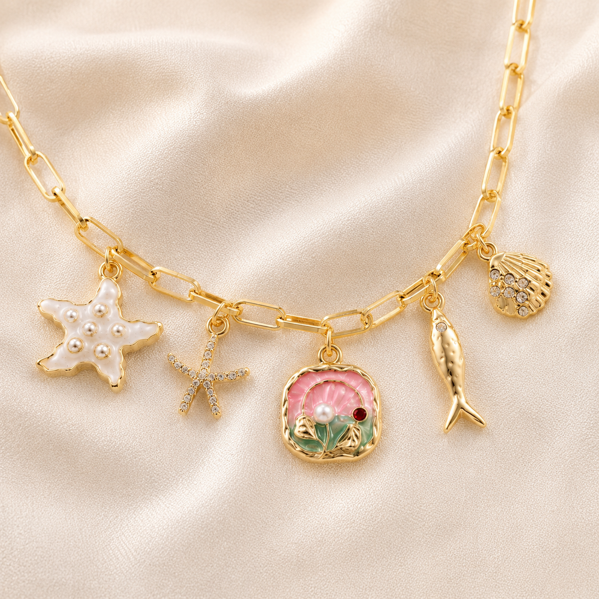 Gold paperclip chain necklace with sea-themed charms including a white pearl starfish, a pink and green clam shell charm, a gold fish charm, and a rhinestone-embellished shell charm, displayed on a beige satin fabric background.