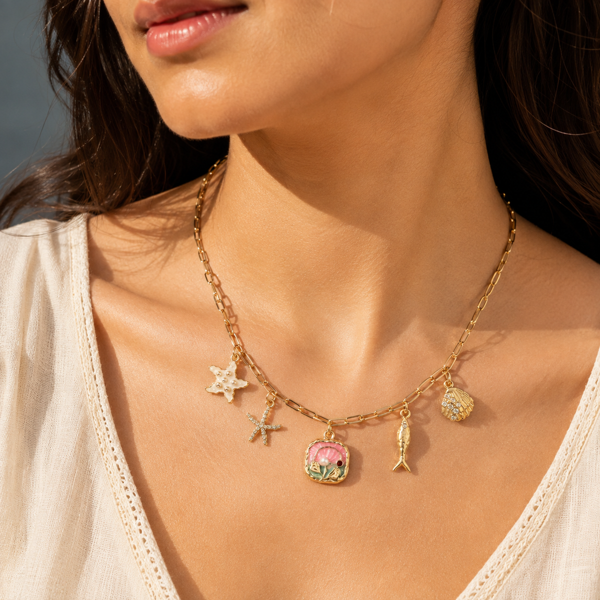 A close-up lifestyle shot of a model wearing a gold chain necklace with various colorful charms, including a pink seashell, starfish, and fish, set against a warm, sunny outdoor background.